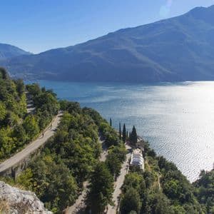 Una strada tortuosa su un verde versante di montagna domina un grande lago soleggiato e lontane catene montuose.
