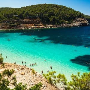 Una vista dall'alto di una cala dove le persone nuotano in acque turchesi poco profonde, con una spiaggia e una scogliera boscosa sullo sfondo.