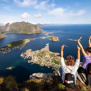 A WeRoad group trip of three women with their backs to the camera sit on a clifftop, overlooking a coastal town, mountains, and a fjord.