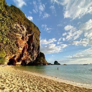 Eine große Felsklippe mit einer Höhle grenzt an einen Sandstrand, an dem Menschen unter einem teilweise bewölkten Himmel schwimmen und entspannen.