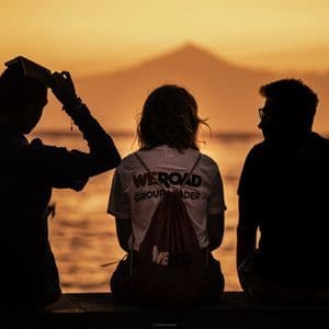Trois personnes d'un voyage en groupe WeRoad sont assises en silhouette sur une jetée, contemplant un coucher de soleil orangé sur l'eau avec une montagne au loin.