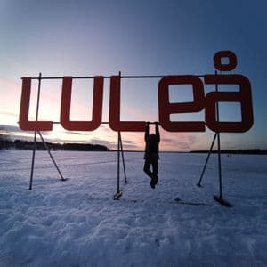 Una persona in controluce appesa alla grande insegna rossa 'LULEÅ' in un paesaggio innevato al tramonto.