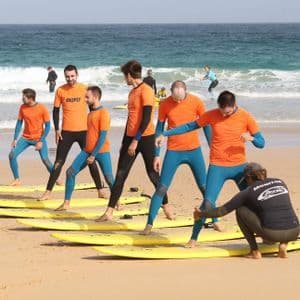 Eine WeRoad-Gruppe in Neoprenanzügen lernt an einem Sandstrand mit gelben Surfbrettern das Surfen von einem Surflehrer.
