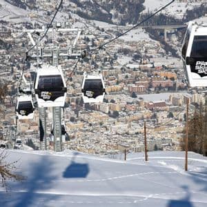 Una fila di bianche funivie sale su un pendio montuoso innevato, affacciandosi su una città nella valle sottostante.