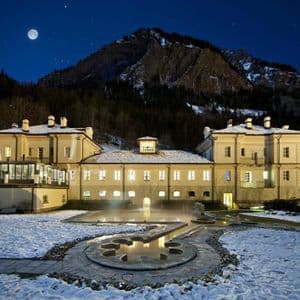 Un edificio illuminato con una piscina termale esterna in un paesaggio innevato, con una grande montagna e una luna piena nel cielo notturno.