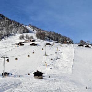 Una località sciistica soleggiata con cabinovie che si muovono su piste innevate, sciatori e chalet in legno sul fianco della montagna.