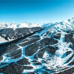 Una vista aerea di una stazione sciistica con diverse piste che attraversano una foresta montana innevata sotto un cielo sereno.