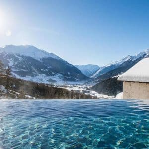 Una piscina a sfioro si affaccia su una valle assolata e montagne innevate, con un tetto innevato in primo piano.
