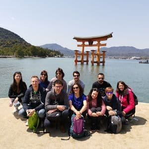 A WeRoad group trip posing for a photo on a shoreline in front of a large orange torii gate standing in the water.