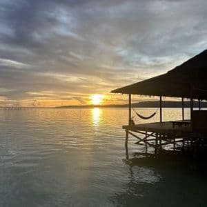 Una hamaca cuelga de una cabaña de madera sobre el agua en calma mientras el sol se pone en el horizonte bajo un cielo nublado.