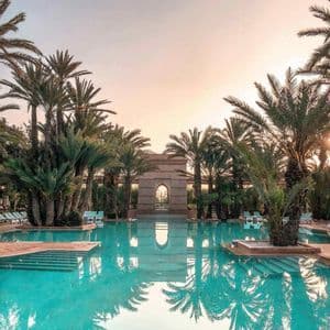 Una grande piscina riflette le palme circostanti e un arco in pietra sotto un cielo al tramonto.