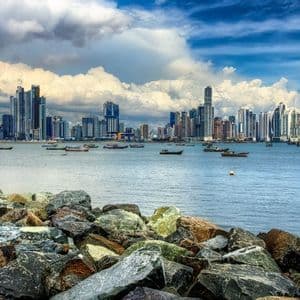 Uno skyline cittadino moderno visto da una costa rocciosa, con piccole imbarcazioni che galleggiano sull'acqua sotto un cielo parzialmente nuvoloso.