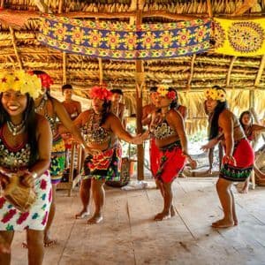 Un gruppo di donne indigene che indossano abiti tradizionali colorati e corone di fiori balla all'interno di una capanna con il tetto di paglia.