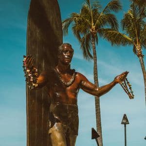 Eine Bronzestatue eines Surfers mit Leis steht neben einem Surfbrett, mit Palmen und dem Ozean im Hintergrund.