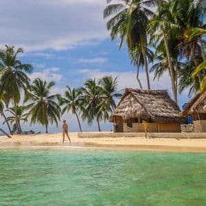Una persona cammina su una spiaggia di sabbia bianca con acqua turchese cristallina, circondata da palme e una capanna di paglia sotto un cielo blu.