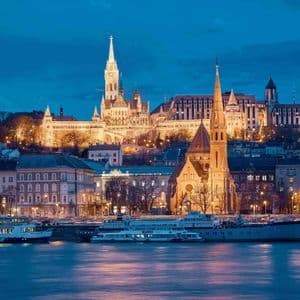 Eine historische europäische Stadtkulisse mit beleuchteten Kirchen und Gebäuden, von einem Flussufer bei Dämmerung aus gesehen.
