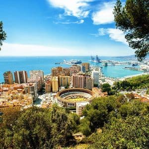 An elevated view of a coastal city featuring a large circular bullring, a commercial port, and the blue sea under a sunny sky.