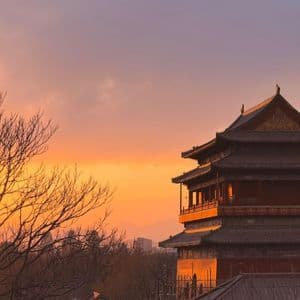 Una tradizionale pagoda a più piani è illuminata dalla luce dorata del tramonto, con alberi spogli e uno skyline cittadino sullo sfondo.