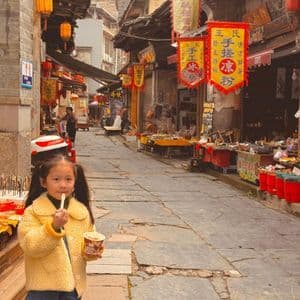 Una ragazza con una giacca in pile gialla mangia da una coppetta mentre si trova su una strada acciottolata in un mercato tradizionale.