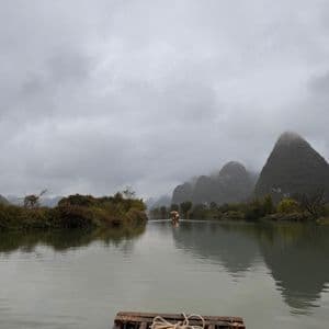 La parte anteriore di una zattera di bambù con corda arrotolata galleggia su un fiume calmo verso montagne nebbiose sotto un cielo coperto.