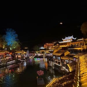 Una cittadina lungo il fiume con architettura tradizionale, illuminata di notte da luci dorate e colorate che si riflettono nell'acqua.