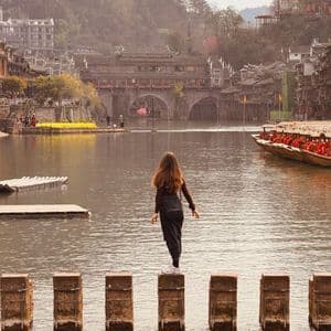 Una donna di spalle alla fotocamera si bilancia su pietre a guado su un fiume, con un villaggio tradizionale e un ponte in lontananza.