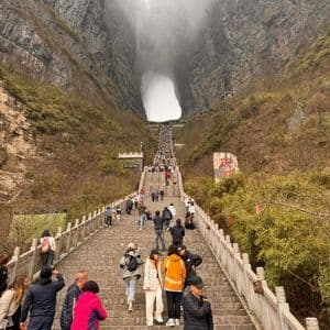 Un gruppo WeRoad in viaggio, con persone che salgono una ripida scalinata in pietra verso un grande arco naturale in una montagna nebbiosa.