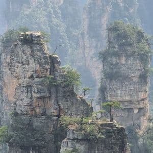Formazioni rocciose alte, simili a pilastri, con alberi verdi in cima, che si ergono attraverso un paesaggio montano nebbioso.