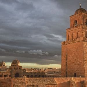 Un minarete de ladrillo se eleva sobre los tejados y cúpulas de la ciudad, iluminado por una luz cálida bajo un cielo nublado y dramático.