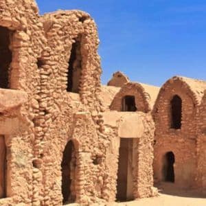 Edificios de piedra desgastados, de color arena, con puertas y ventanas arqueadas, bajo un cielo azul claro.