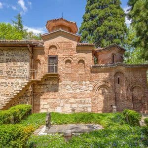 Un'antica chiesa in pietra e mattoni rossi, circondata da rigogliosi alberi verdi e un piccolo giardino con una tomba.