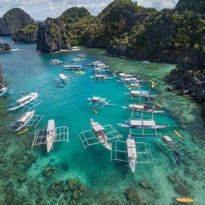 An aerial view of numerous outrigger boats and kayaks floating in a clear turquoise lagoon surrounded by steep, jungle-covered cliffs.