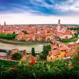 Un ampio paesaggio urbano di edifici dai tetti rossi lungo un fiume sinuoso con un ponte ad arco in pietra, visto da una collina con fiori rossi.