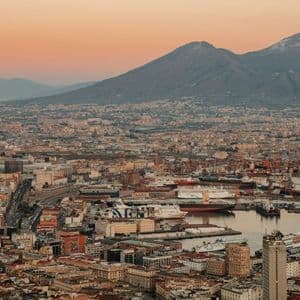 Una vista aérea de una extensa ciudad costera y su concurrido puerto al pie de un gran volcán bajo un cielo de atardecer de tonos cálidos.