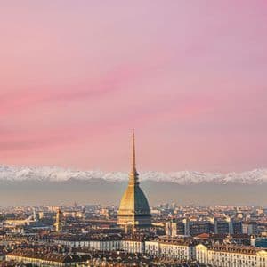 Una vista aerea di una città con un edificio centrale a cupola, sullo sfondo di montagne innevate sotto un cielo rosa al tramonto.