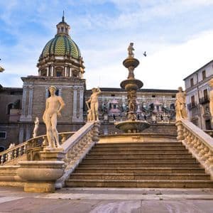 Una fontana riccamente ornata e un'ampia scalinata fiancheggiata da statue classiche nude in una piazza storica sotto un cielo azzurro.