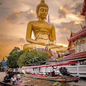 Des bateaux à longue queue naviguent sur un canal, avec une statue géante de Bouddha doré et un temple en arrière-plan, au coucher du soleil.
