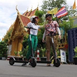Une femme ajuste le casque d'un homme alors qu'ils se tiennent avec des trottinettes électriques devant un temple doré orné d'un toit de tuiles rouges.