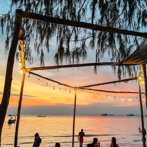 Un groupe WeRoad se détend dans un bar de plage sous des guirlandes lumineuses, regardant le coucher de soleil sur la mer.
