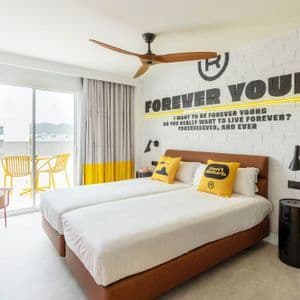A modern hotel room with two single beds, yellow accent pillows, and a "Forever Young" mural on a white brick wall.