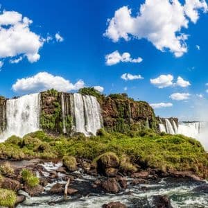 Ein Panoramablick auf ein riesiges Wasserfallsystem, das über felsige Klippen mit grüner Vegetation unter einem strahlend blauen, bewölkten Himmel herabstürzt.