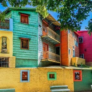 Exterior view of stacked, colorful buildings painted turquoise, yellow, red, and blue under green tree leaves and a clear sky.
