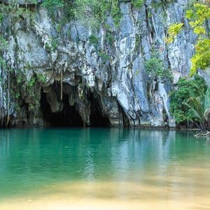 Ein dunkler Höhleneingang am Fuße einer vegetationsbedeckten Felsklippe, der sich zu einem ruhigen, smaragdgrünen Gewässer mit sandigem Ufer öffnet.