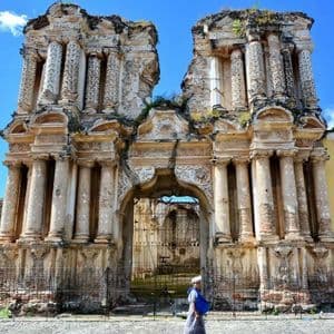 A person in traditional clothing walks on a cobblestone street in front of a large, ornate, and crumbling stone facade.