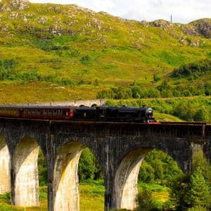 Un treno a vapore nero con carrozze rosse attraversa un viadotto in pietra, con una rigogliosa montagna verde sullo sfondo.