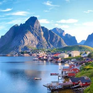 Un villaggio marittimo con case rosse su palafitte si trova su una baia calma ai piedi di imponenti montagne sotto un cielo blu.