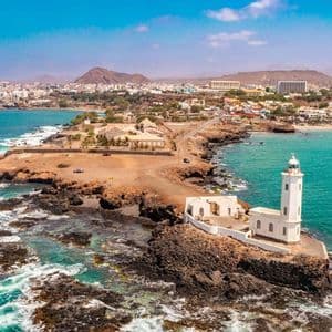 An aerial view of a white lighthouse on a rocky peninsula, with a coastal city and mountains in the background under a blue sky.