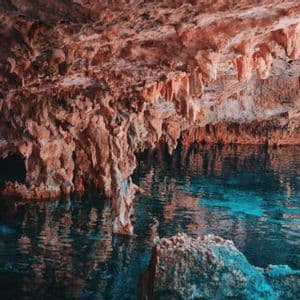 Reddish-orange stalactites hang from the ceiling of a cave above a pool of clear, turquoise water.