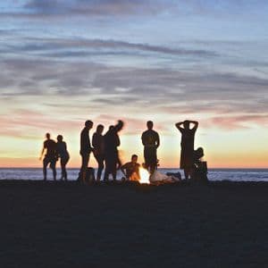 Silhouettes of a WeRoad group trip gathered around a bonfire on a beach at sunset, with the ocean in the background.