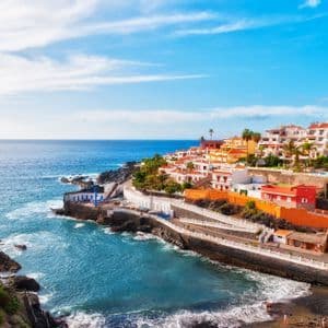 Eine farbenfrohe Küstenstadt mit weißen und orangefarbenen Gebäuden liegt an einer felsigen Küste direkt am blauen Meer unter einem teilweise bewölkten Himmel.